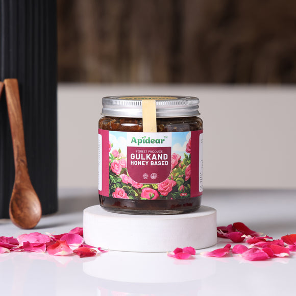 A jar of Apidear Gulkand honey-based spread on a white surface with an array of pink rose petals scattered around it. A wooden spoon is also visible in the background.