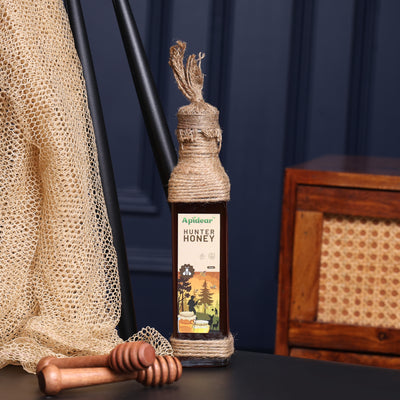 A bottle of Hunter Honey with a honey dipper in front, set on a dark surface with a woven table runner and a wooden object in the background.